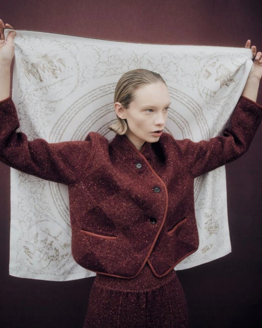 Fashionable woman in a maroon jacket holds a patterned scarf indoors, creating a stylish portrait with a muted backdrop.