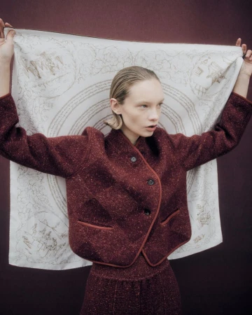 Fashionable woman in a maroon jacket holds a patterned scarf indoors, creating a stylish portrait with a muted backdrop.