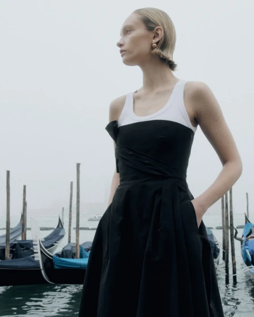 A woman poses near gondolas in a black dress and white tank top in an outdoor setting, with a cloudy sky overhead in Venice, Italy.