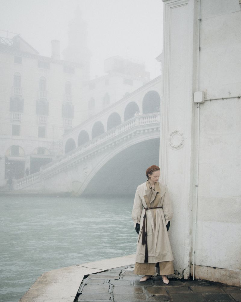 A woman in a beige trench coat stands on a stone edge next to a canal, with a large arched bridge in the background on a foggy day.