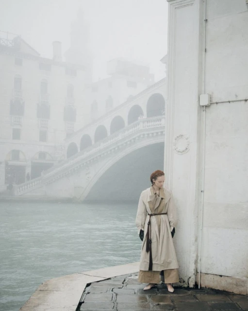 A woman in a beige trench coat stands on a stone edge next to a canal, with a large arched bridge in the background on a foggy day.