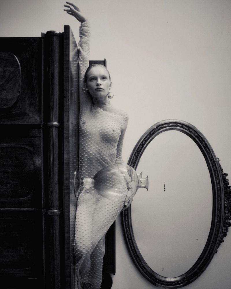 Monochrome fashion portrait of a slim adult woman in sheer polka dot dress, posing next to a vintage wardrobe and oval mirror against a neutral wall.