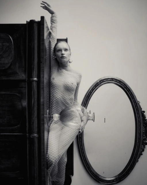 Monochrome fashion portrait of a slim adult woman in sheer polka dot dress, posing next to a vintage wardrobe and oval mirror against a neutral wall.