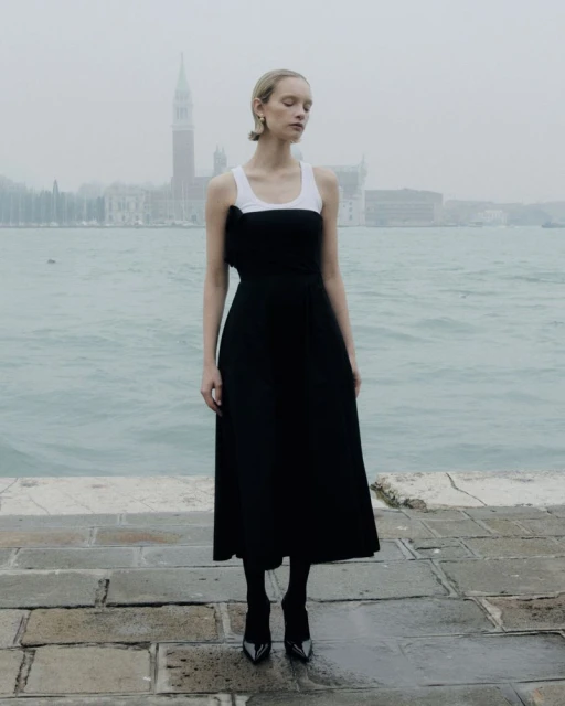 A woman in a black and white dress stands on a stone platform with the ocean and cityscape of Venice, Italy behind her in a fashion editorial.