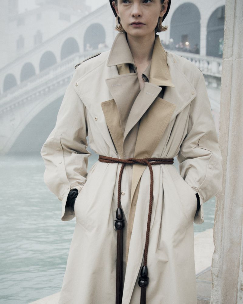 Woman in a beige trench coat with a brown rope belt posing in front of a stone bridge over the water, creating a fashionable, elegant image.