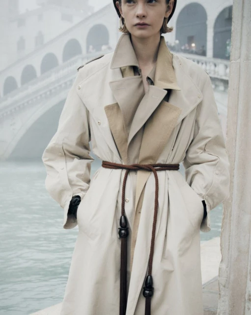 Woman in a beige trench coat with a brown rope belt posing in front of a stone bridge over the water, creating a fashionable, elegant image.
