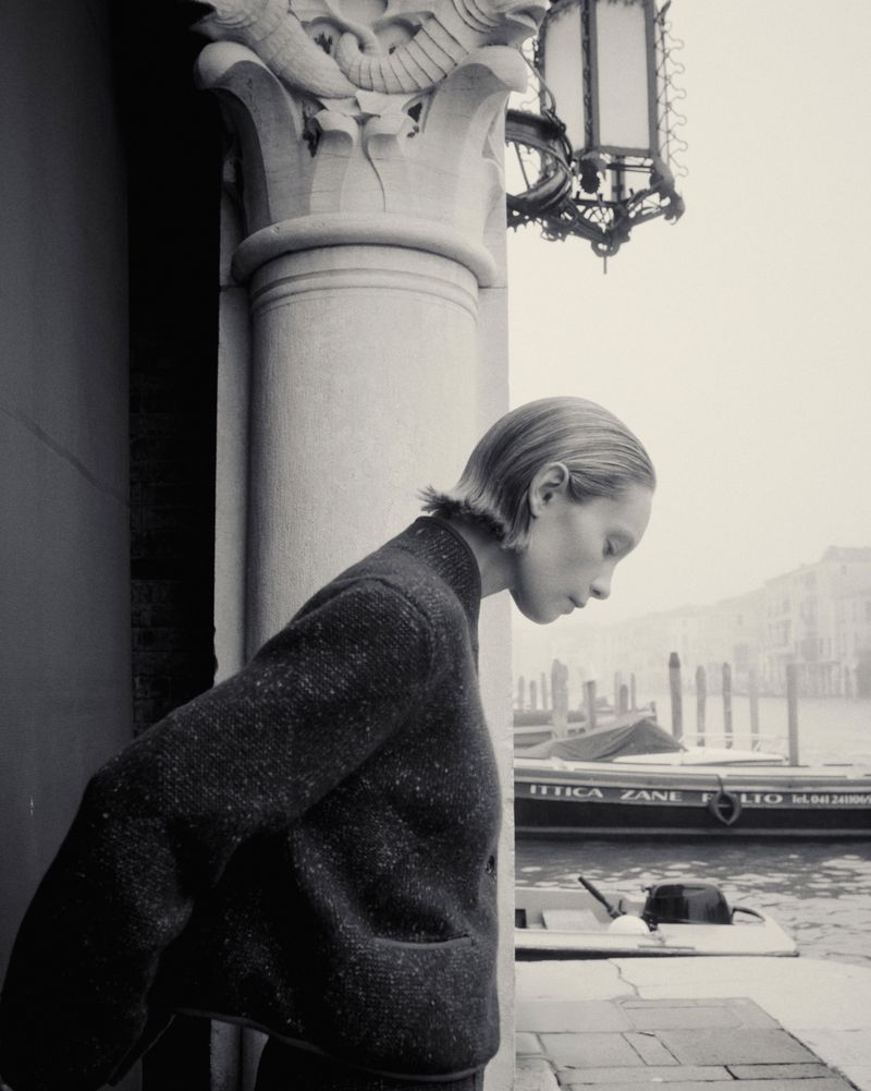 Black and white portrait of a young woman in a sweater standing near a canal in Venice, Italy, with historic buildings in the background.