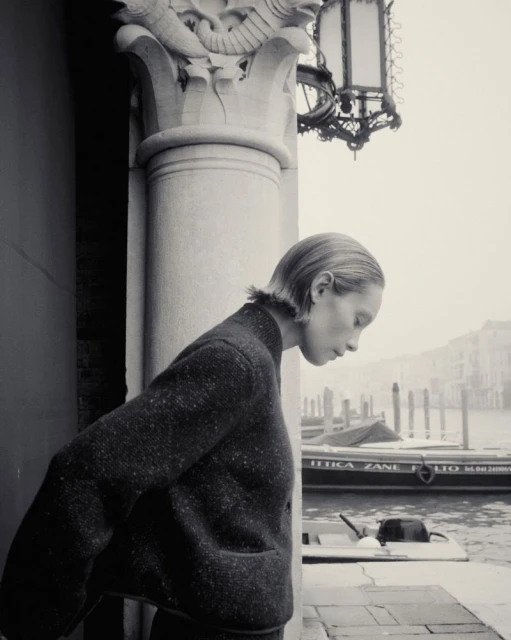 Black and white portrait of a young woman in a sweater standing near a canal in Venice, Italy, with historic buildings in the background.