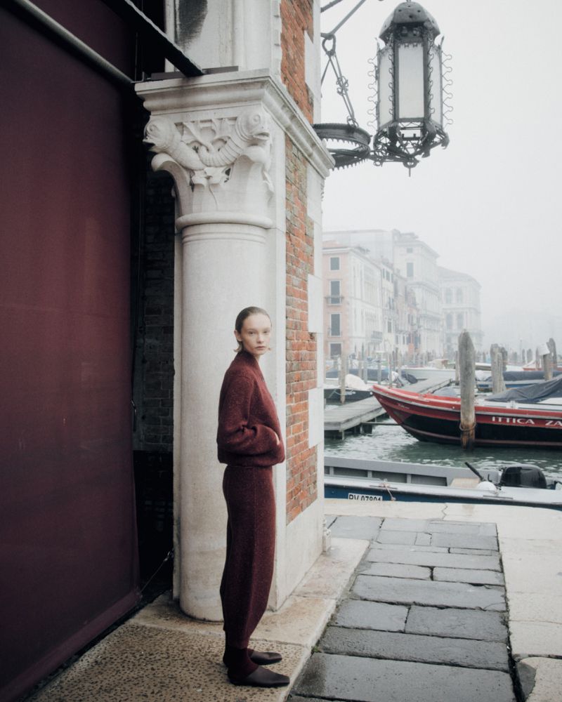 Fashion shot of woman in a knit pantsuit standing near a canal in Venice, Italy on a foggy day, with boats visible in the background.