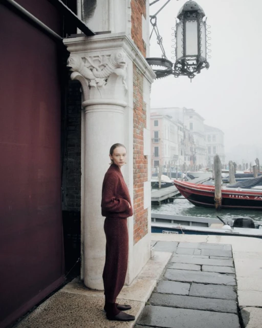 Fashion shot of woman in a knit pantsuit standing near a canal in Venice, Italy on a foggy day, with boats visible in the background.