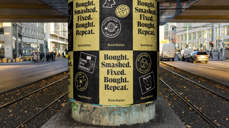 Outdoor advertisement on a pillar with repeating graphics and typography. Urban scene with street traffic and buildings in the background.