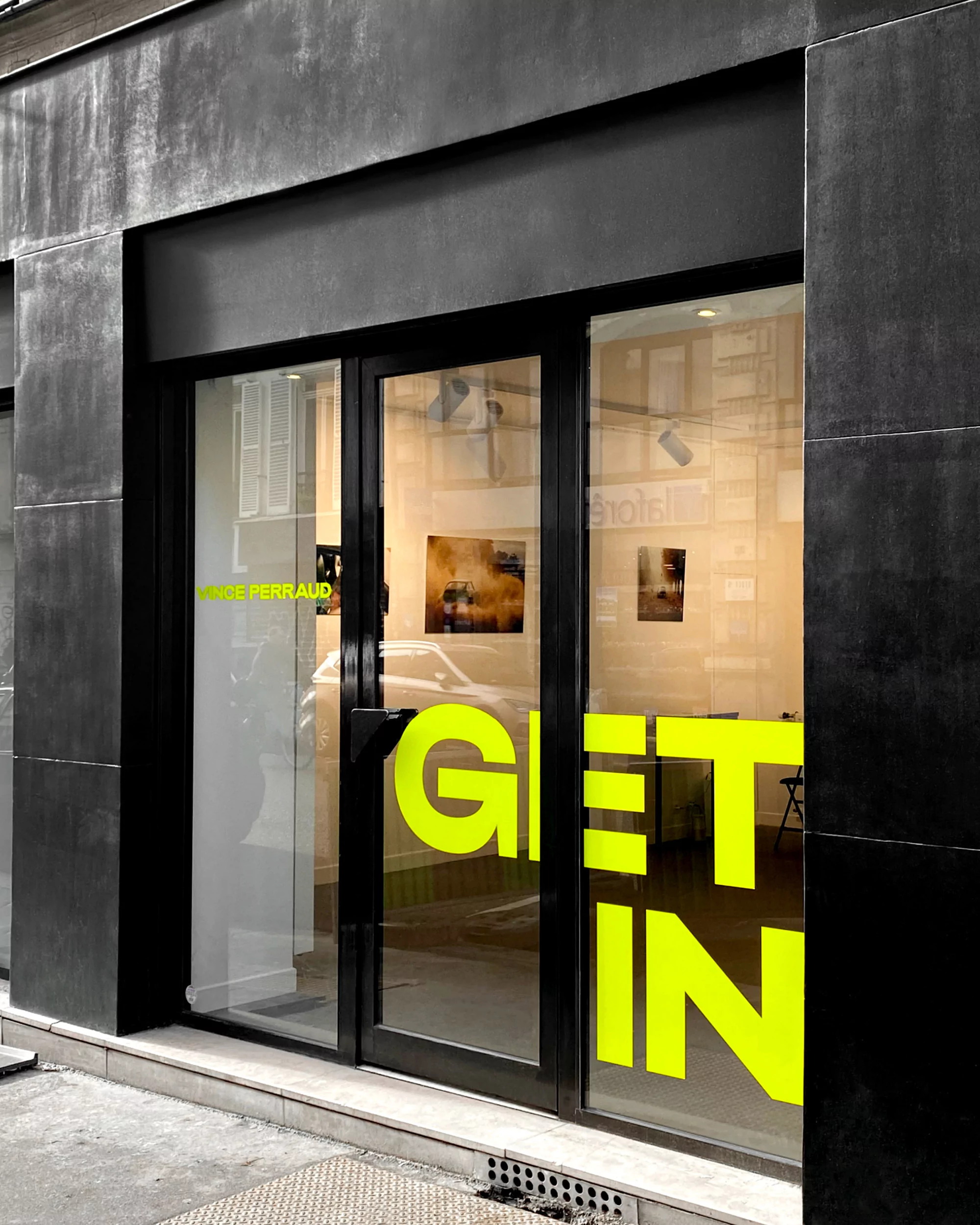 Exterior of a modern art gallery with a sleek black facade and glass doors displaying a vibrant "GET IN" sign and artwork inside.
