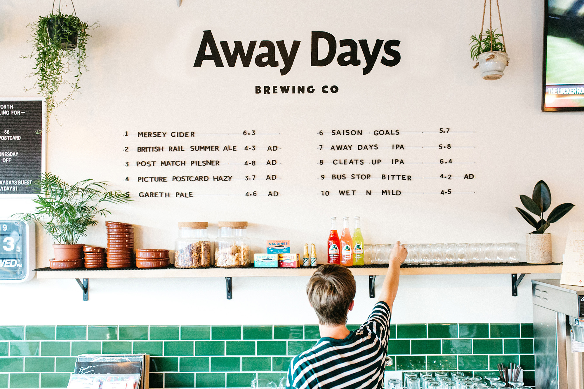 A person reaches for a glass behind a counter inside Away Days Brewing Co. in a bright, naturally-lit interior with green tile.