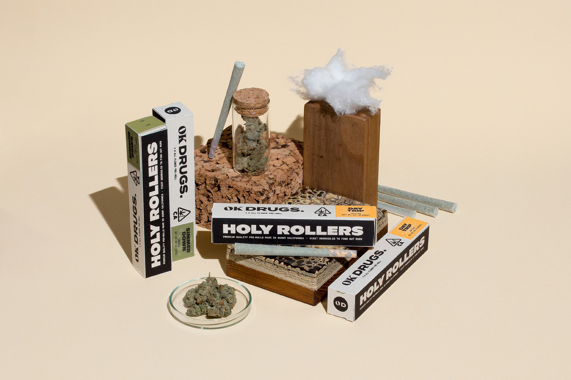 Still life showcasing cannabis product packaging from OK Drugs, featuring 'Holy Rollers' pre-rolls, buds in a dish, and other accessories.