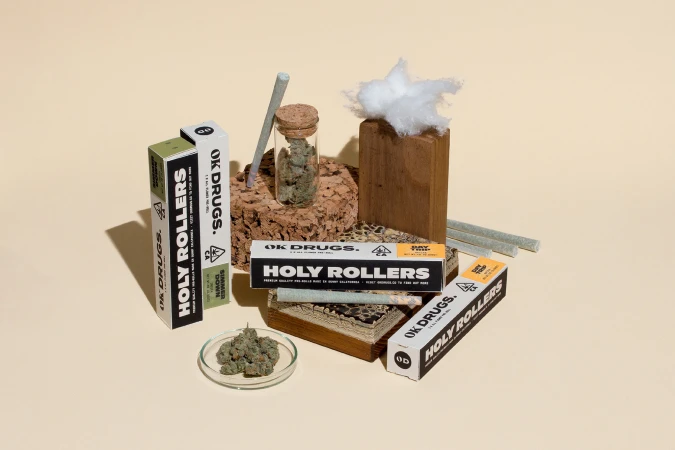 Still life showcasing cannabis product packaging from OK Drugs, featuring 'Holy Rollers' pre-rolls, buds in a dish, and other accessories.