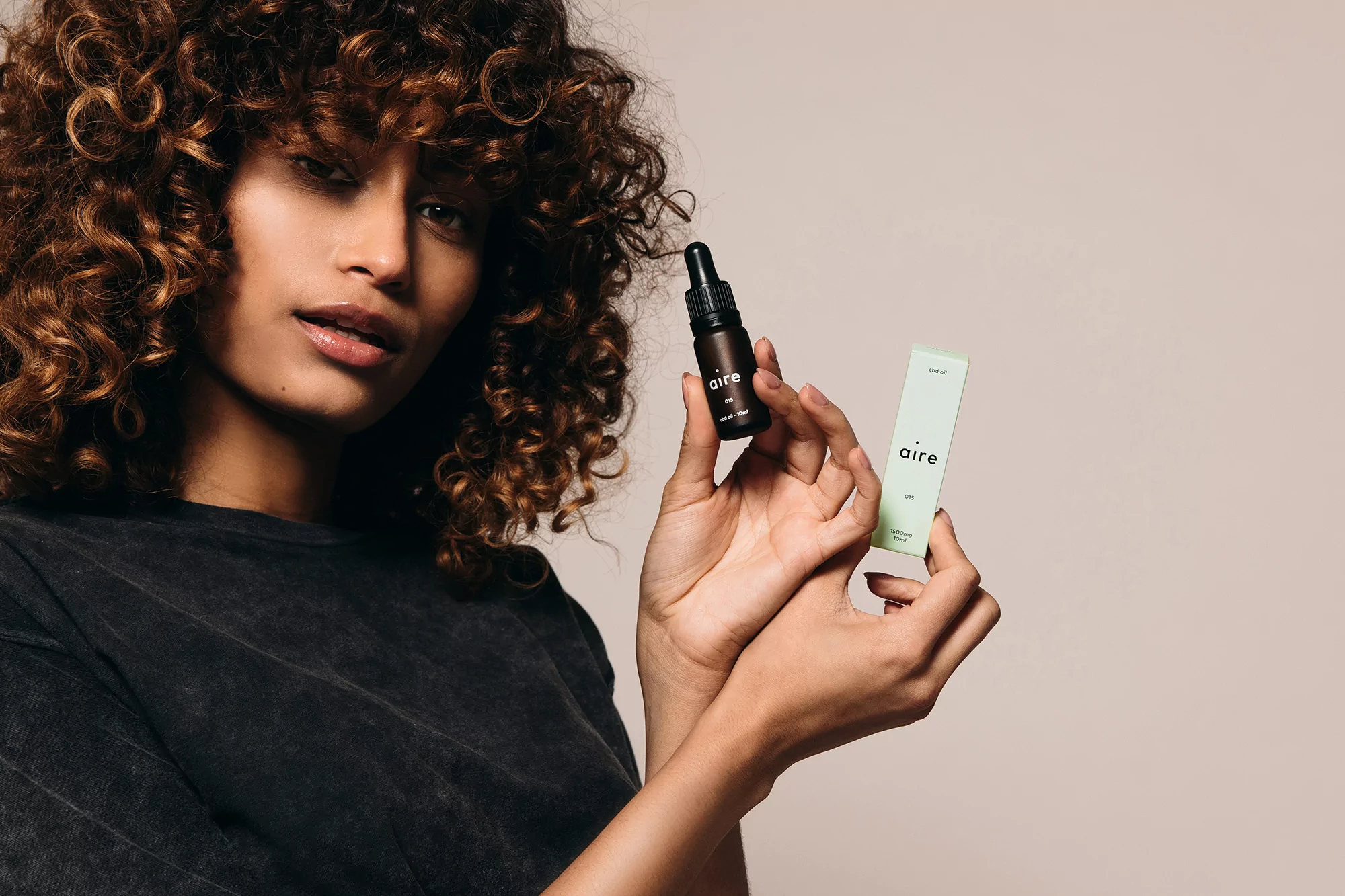 Brunette woman presents small brown bottle and a light green box of skincare or cosmetic product in a studio setting.