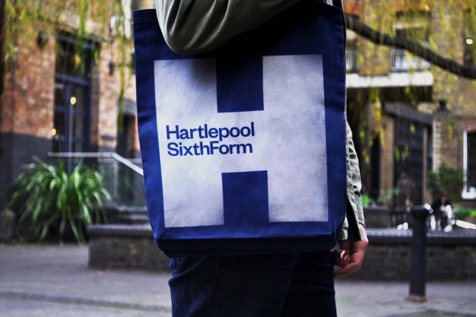 Person holding a blue canvas tote bag with a white H and Hartpool Sixth Form branding outdoors.