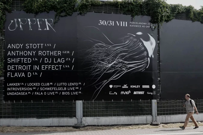 Outdoor view of a music event advertisement with an abstract design and text, featuring a pedestrian in motion on the sidewalk.