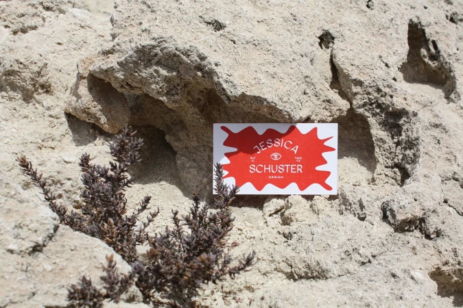 Close-up of a business card for Jessica Schuster Design placed against a rock formation, accented by a small plant. Bright, natural lighting.