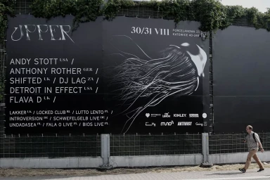 Large black event poster with white typography mounted on a wire fence outdoors with a pedestrian walking past