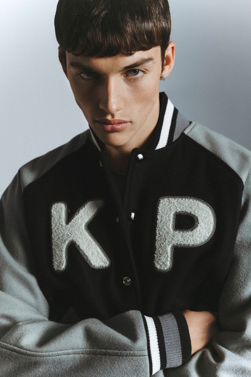 Close-up of a male model wearing a black and grey varsity jacket with white letters 'KP', arms crossed, against a grey gradient backdrop.