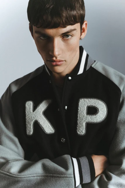 Close-up of a male model wearing a black and grey varsity jacket with white letters 'KP', arms crossed, against a grey gradient backdrop.