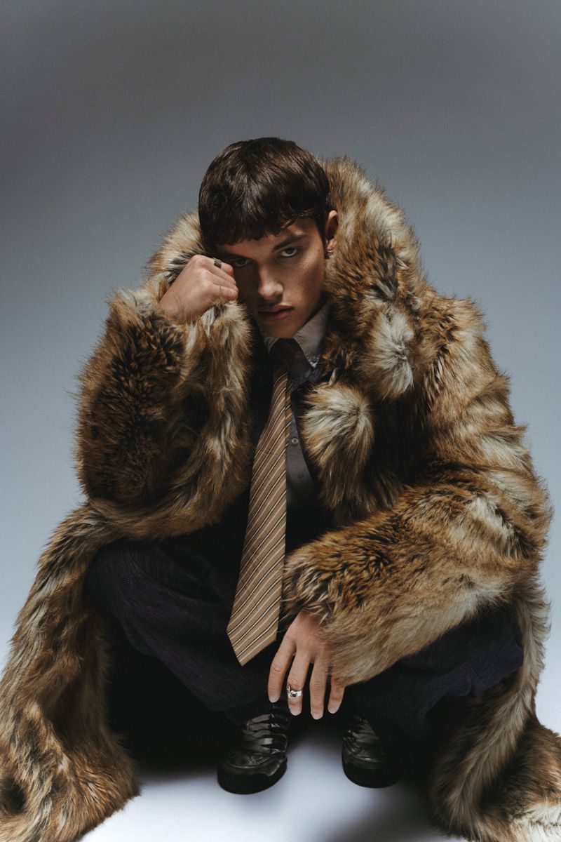 Male model in a fur coat crouching in a studio setting. He is wearing a tie and dress shoes, looking directly at the camera.