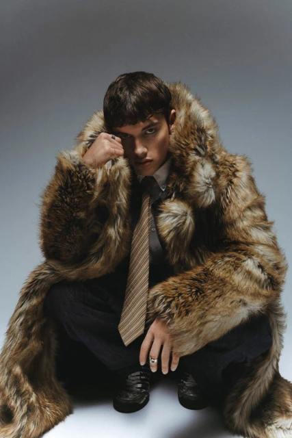 Male model in a fur coat crouching in a studio setting. He is wearing a tie and dress shoes, looking directly at the camera.