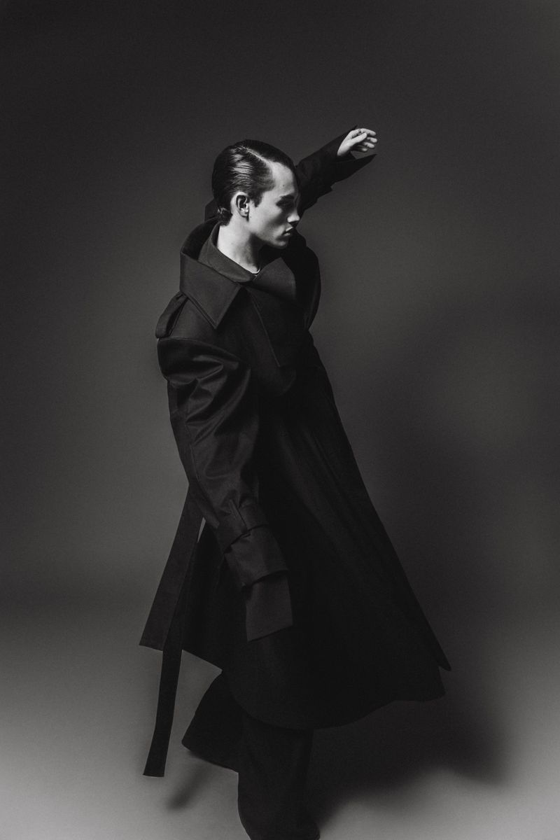 A stylish man in a dramatic black coat strikes a pose against a grey background, emphasizing fashion and monochrome aesthetics.