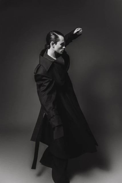 A stylish man in a dramatic black coat strikes a pose against a grey background, emphasizing fashion and monochrome aesthetics.