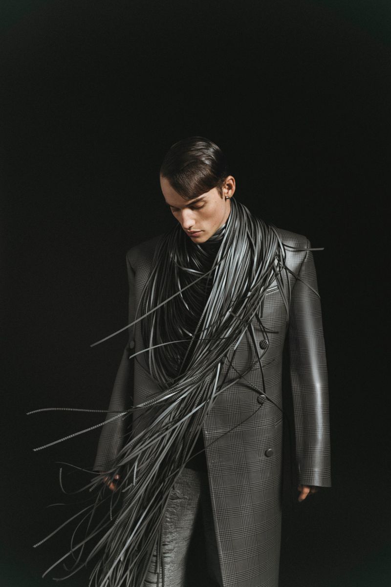 Fashion shot of a slender male model wearing a grey coat and pants, adorned with a striking arrangement of black cables.