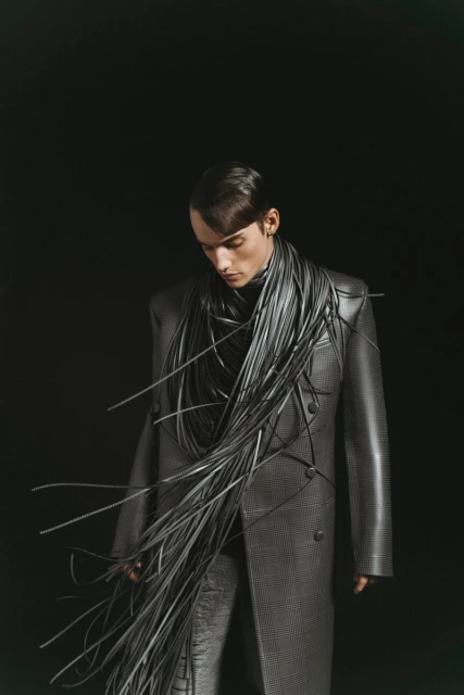 Fashion shot of a slender male model wearing a grey coat and pants, adorned with a striking arrangement of black cables.