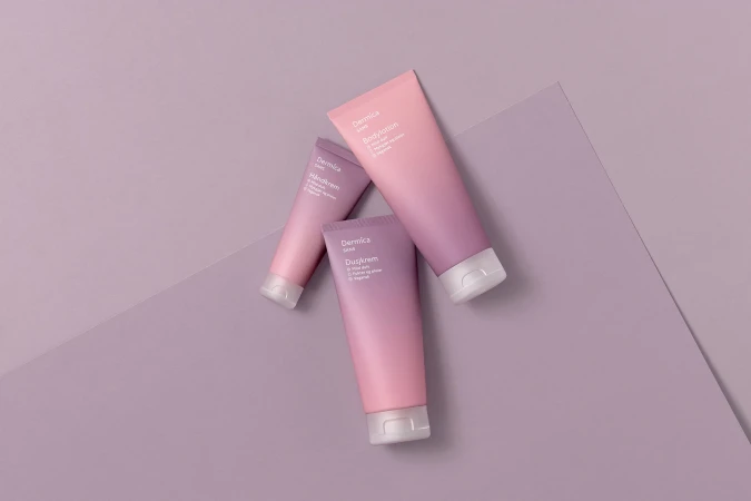 Three Dermica Sans body lotion and hand cream tubes in shades of pink and purple are displayed on a light purple surface.