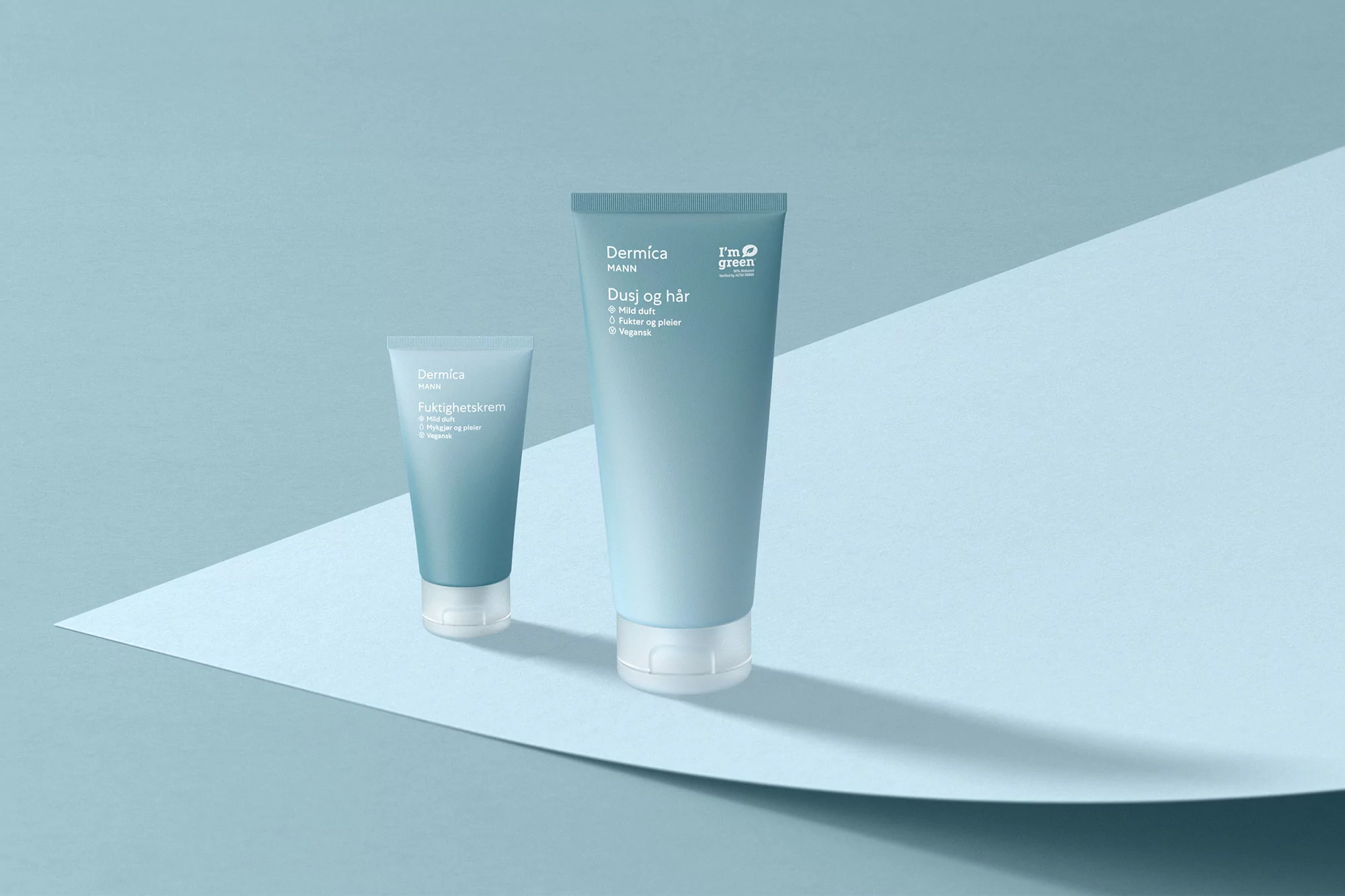Two light blue Dermica MANN skincare tubes, one large and one small, sit on a light blue surface with a folded backdrop.