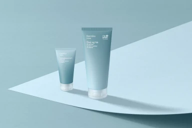 Two blue Dermica skincare tubes standing upright on a curved pastel blue paper backdrop studio set
