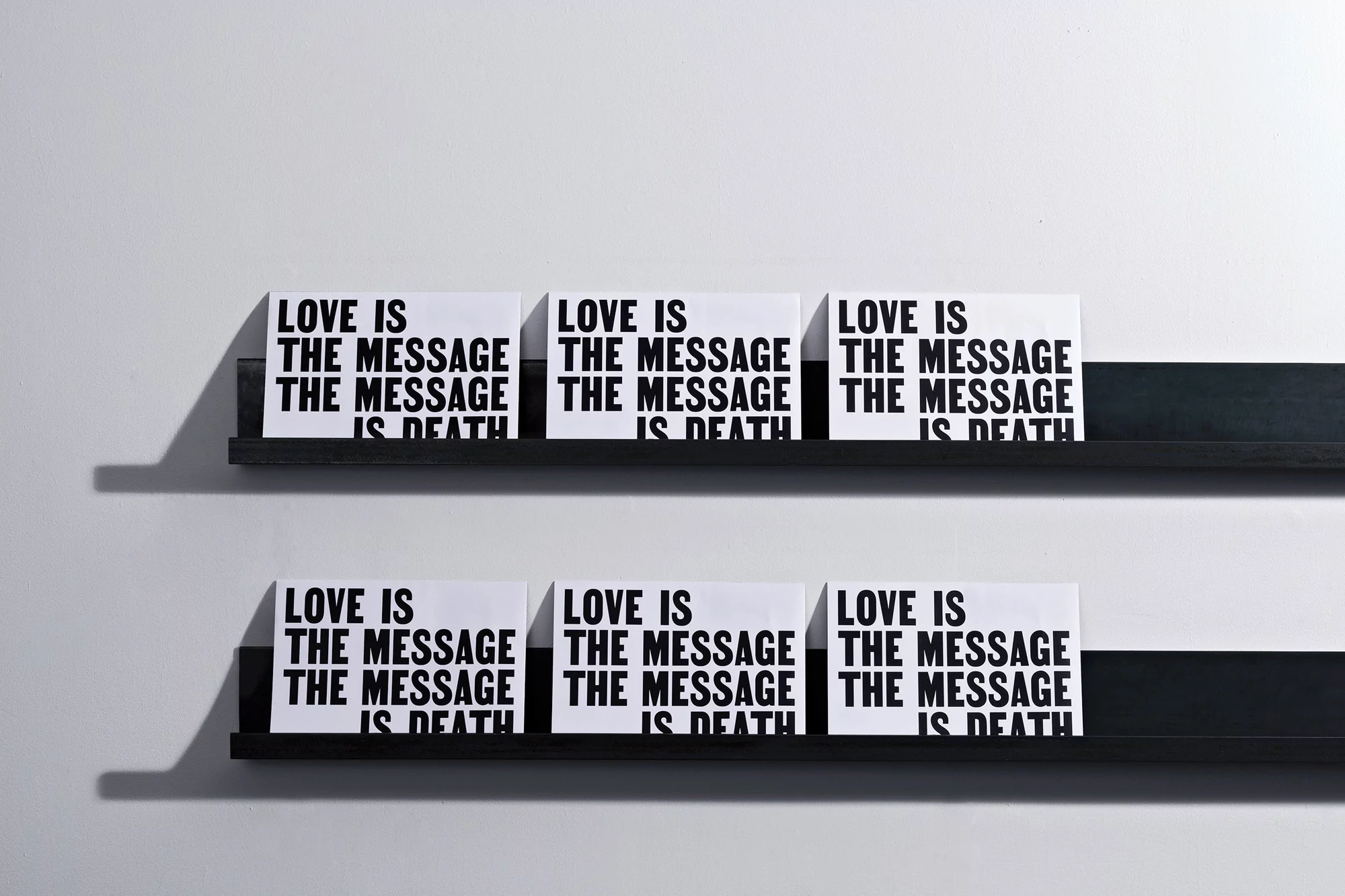 Six white signs with black text reading "Love is the message is death" are displayed on two black shelves against a white wall.