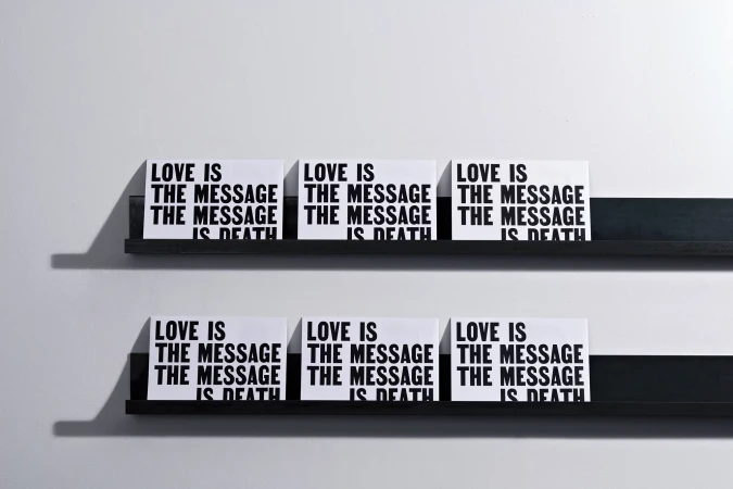 Six white signs with black text reading "Love is the message is death" are displayed on two black shelves against a white wall.