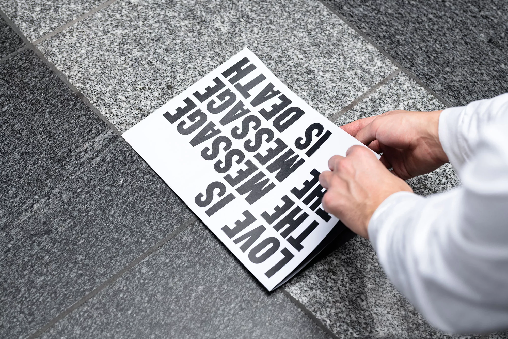 A person holds a paper with the text 'LOVE IS THE MESSAGE' in black letters on a gray tiled floor, creating a thought-provoking visual statement.