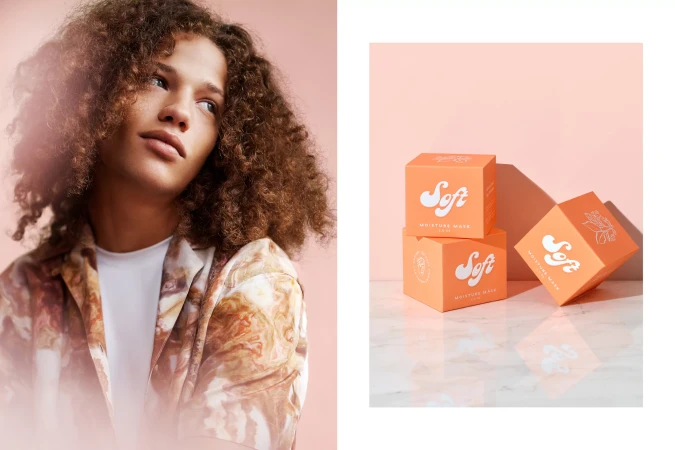 A young adult with curly hair gazes left, standing next to stacked peach-colored skincare boxes on a light marble surface.