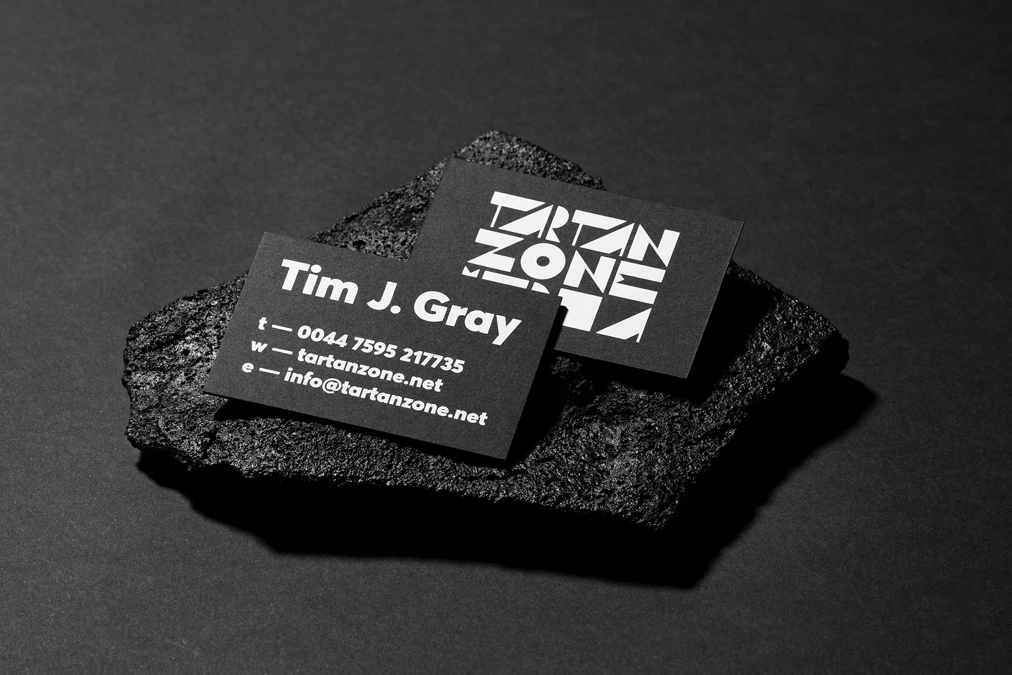 Two black business cards with white text are displayed on a textured black rock, set against a dark background, providing a minimalist aesthetic.