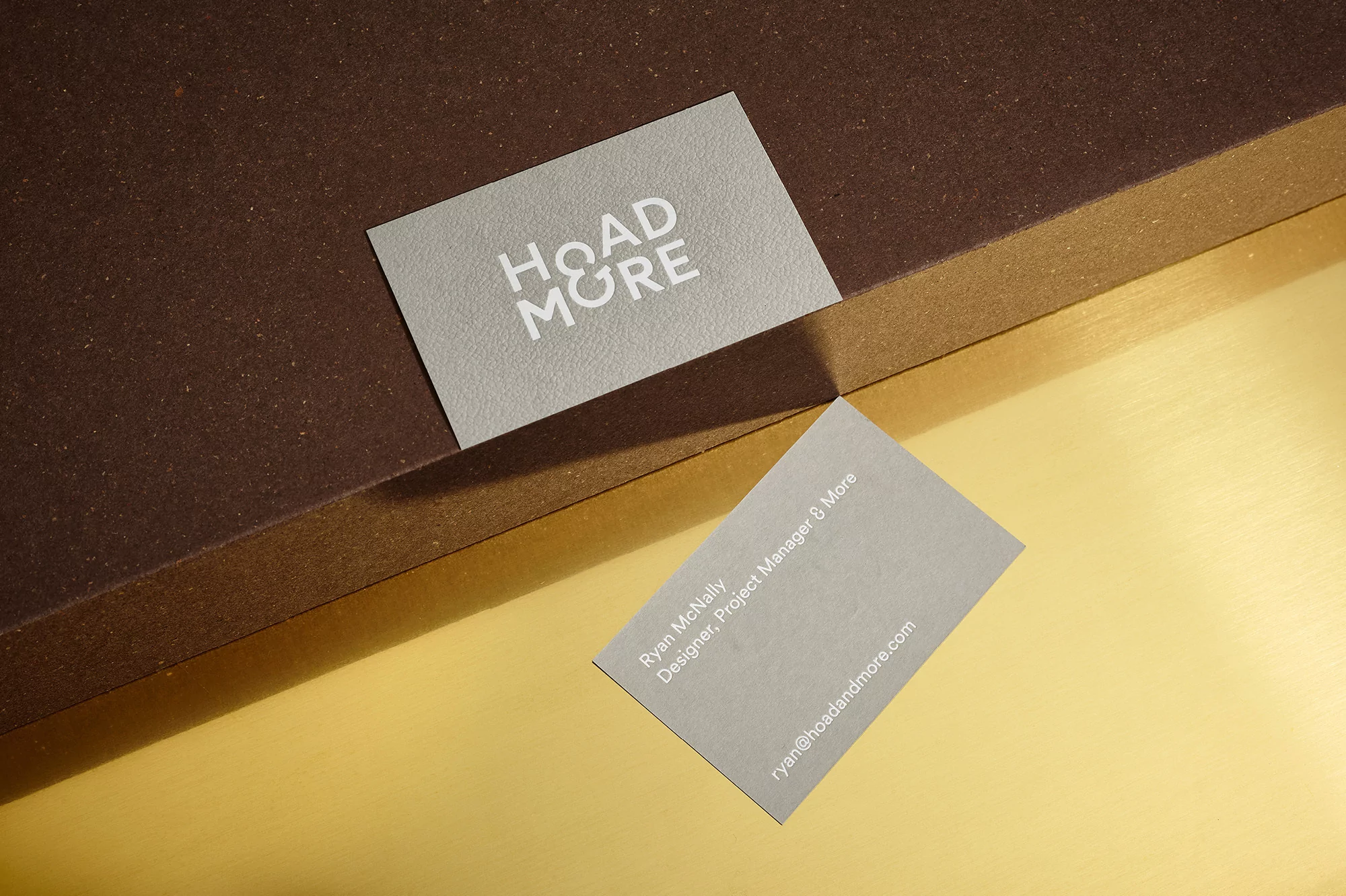 Two gray business cards displaying 'HOAD & MORE' on a layered surface of brown and gold with diffused lighting, highlighting minimalist design.