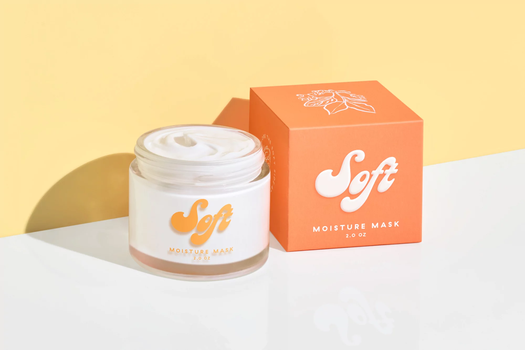 Product shot of a jar and box of "Soft" moisture mask against a yellow backdrop, emphasizing skincare product presentation.
