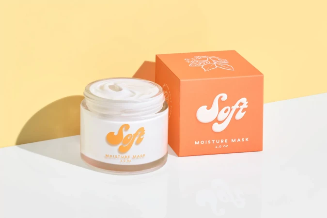 Product shot of a jar and box of "Soft" moisture mask against a yellow backdrop, emphasizing skincare product presentation.