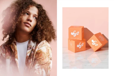 Portrait of young person with curly brown hair beside orange product boxes on a reflective marble surface