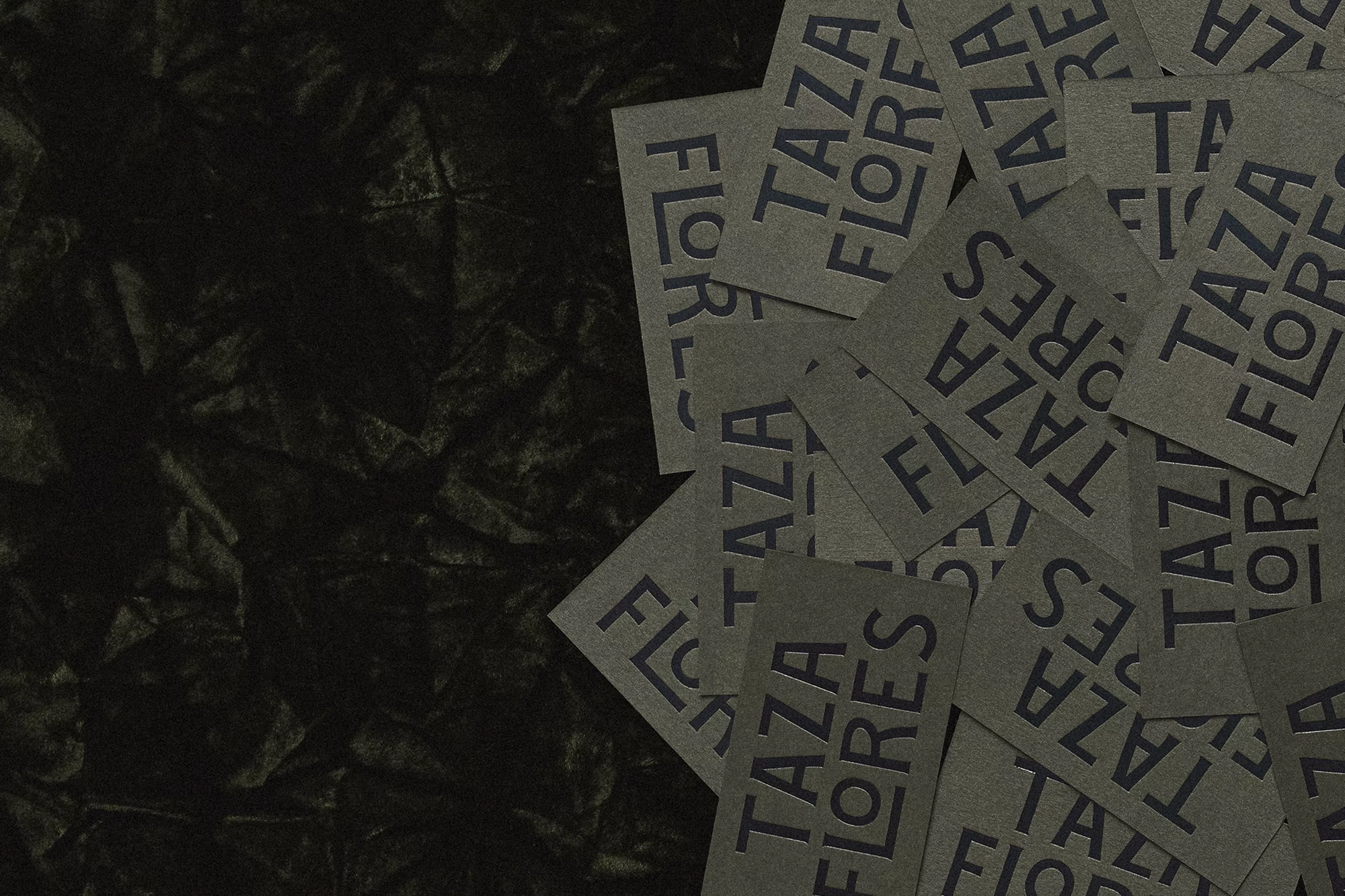 Close-up of gray cards with the text 'Taza Flores' arranged on a dark gray wrinkled background with a shallow depth of field.