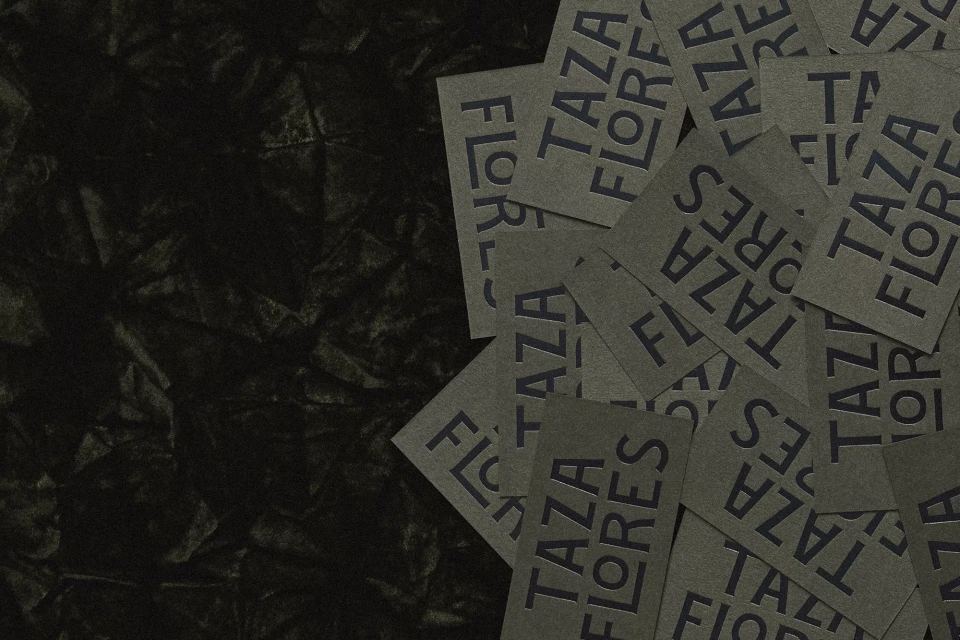 Close-up of gray cards with the text 'Taza Flores' arranged on a dark gray wrinkled background with a shallow depth of field.