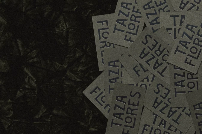 Close-up of gray cards with the text 'Taza Flores' arranged on a dark gray wrinkled background with a shallow depth of field.