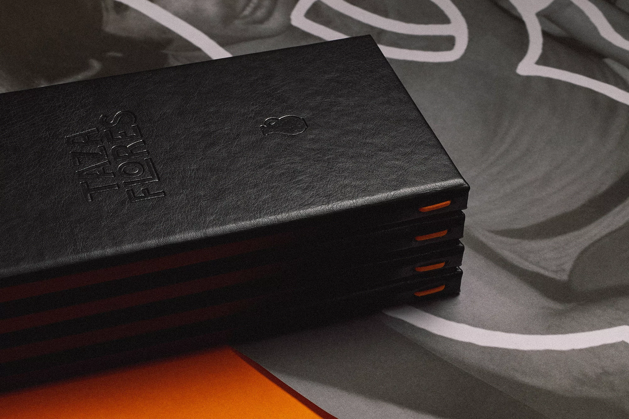 Stack of black journals with orange binding accents resting on a gray surface with white patterned graphics, clean product photography.