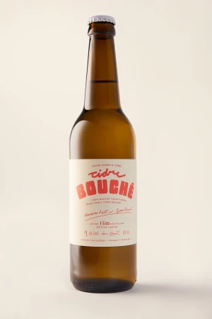 Studio shot of a brown glass cider bottle featuring a white label with red text against a neutral backdrop. Food and beverage.