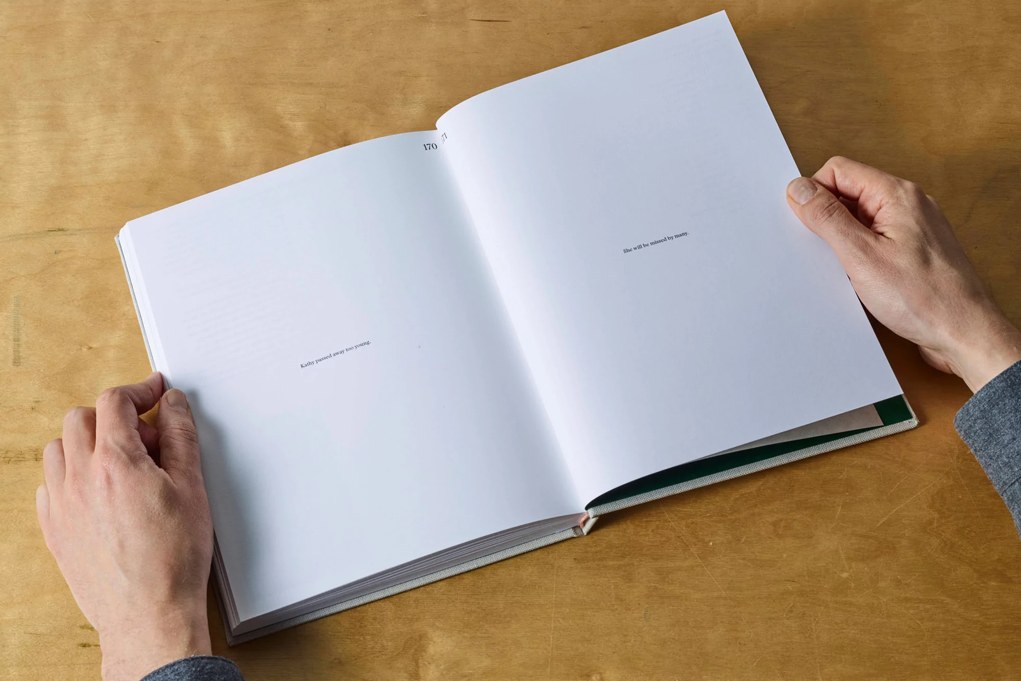 Open book being held by a person with pages displaying text, set on a light brown wooden table indoors in natural light.